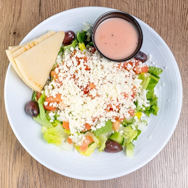 A fresh Greek salad with feta, olives, tomatoes, lettuce, pita bread, and dressing on a white plate.
