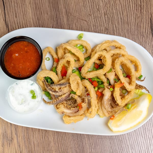Crispy fried calamari with red sauce, white sauce, lemon wedge, and chopped greens on a white plate.